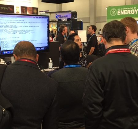 A group stands at the DOE booth watching a demonstration.